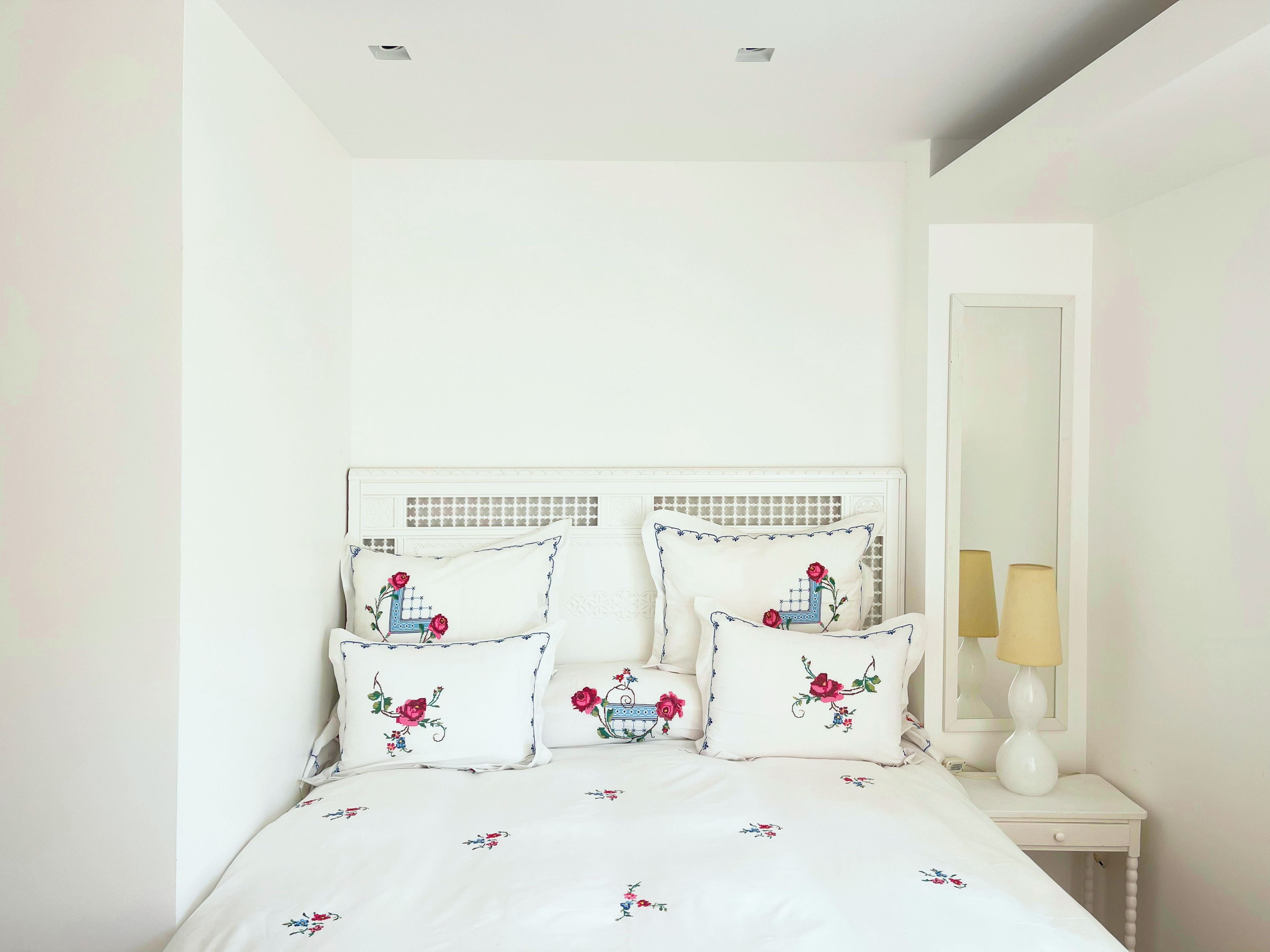 Een bed met bloemenbedekking en decoratieve kussens, een nachtkastje met een lamp, en een spiegel aan de muur.