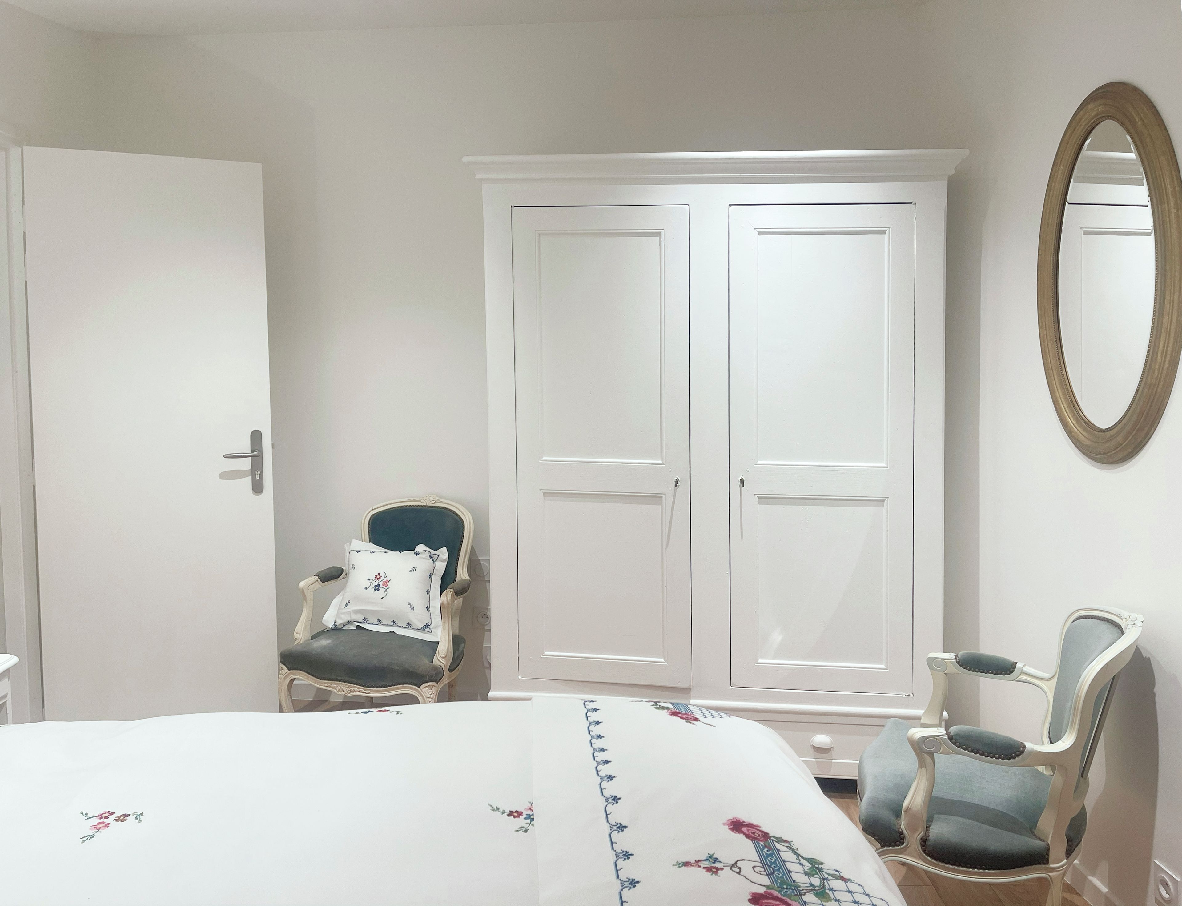 A bedroom with a bed, two chairs, a wardrobe, and a mirror.