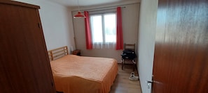 Un dormitorio con cama, una silla, un escritorio, una ventana con cortinas y una puerta.
