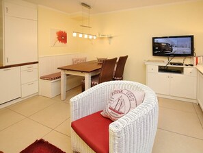 Un espace de vie compact comprenant une table à manger, des meubles en osier blancs, un téléviseur et un coussin rouge.