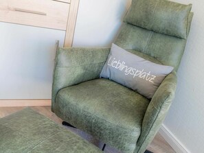 Un fauteuil vert recouvert de tissu, agrémenté d’un coussin sur lequel est inscrit le mot « Lieblingsplatz », dans une pièce dotée d’un plancher en bois et d’un mur de couleur claire.