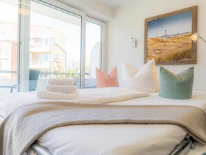 A hotel room with a bed, pillows, a painting, and a window with a view of buildings. - Holiday Apartment With 2 Bedrooms (St. Peter-Ording)