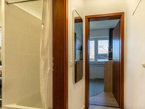 A bathroom with a shower, a wooden door leading to a hallway, and a window with a view of the outside. - 1 in the Westmarken Building (St. Peter-Ording)