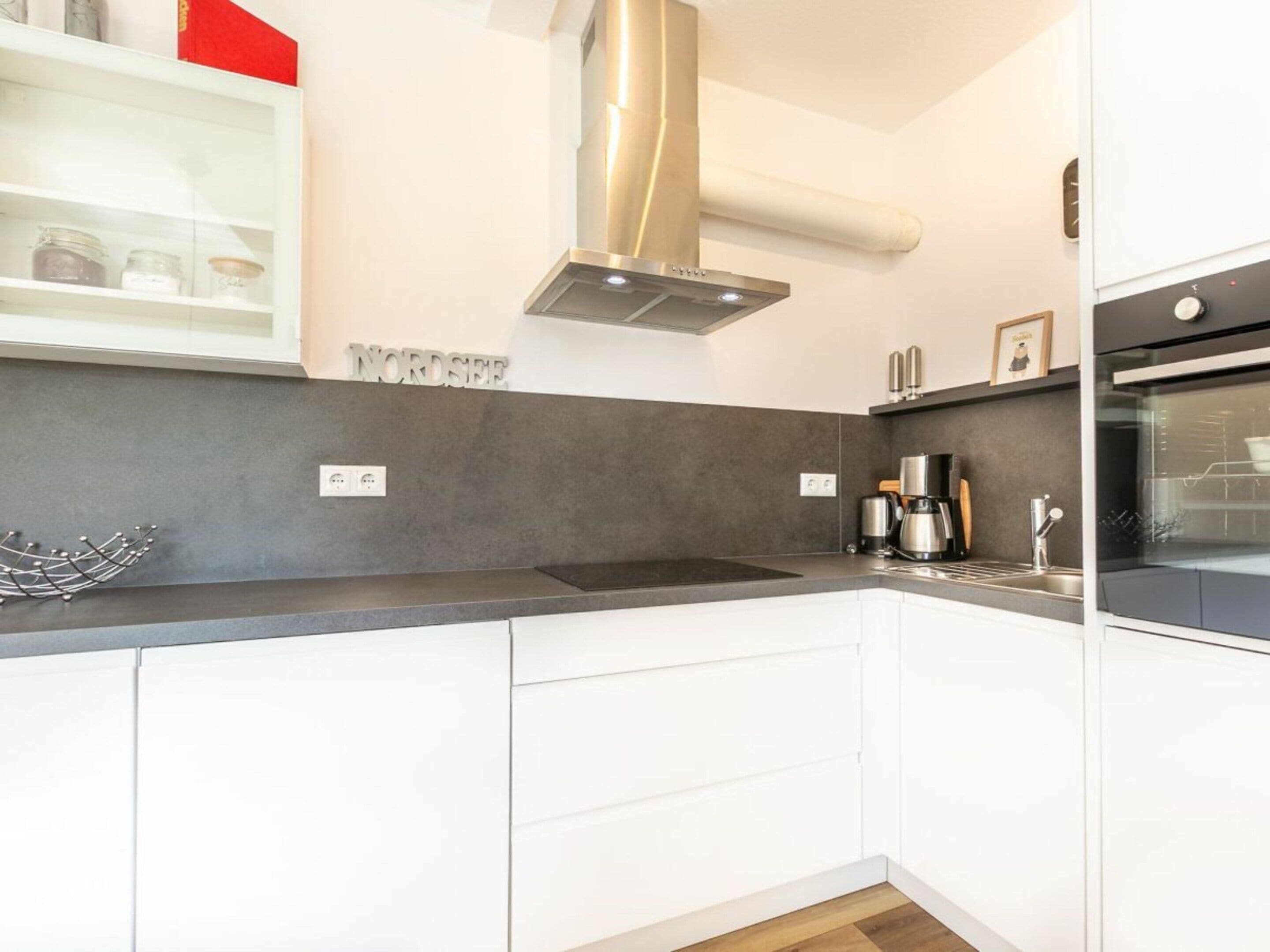 Une cuisine moderne avec des armoires blanches, une hotte de cuisinière en acier inoxydable, un four encastré et une machine à café posée sur le plan de travail.