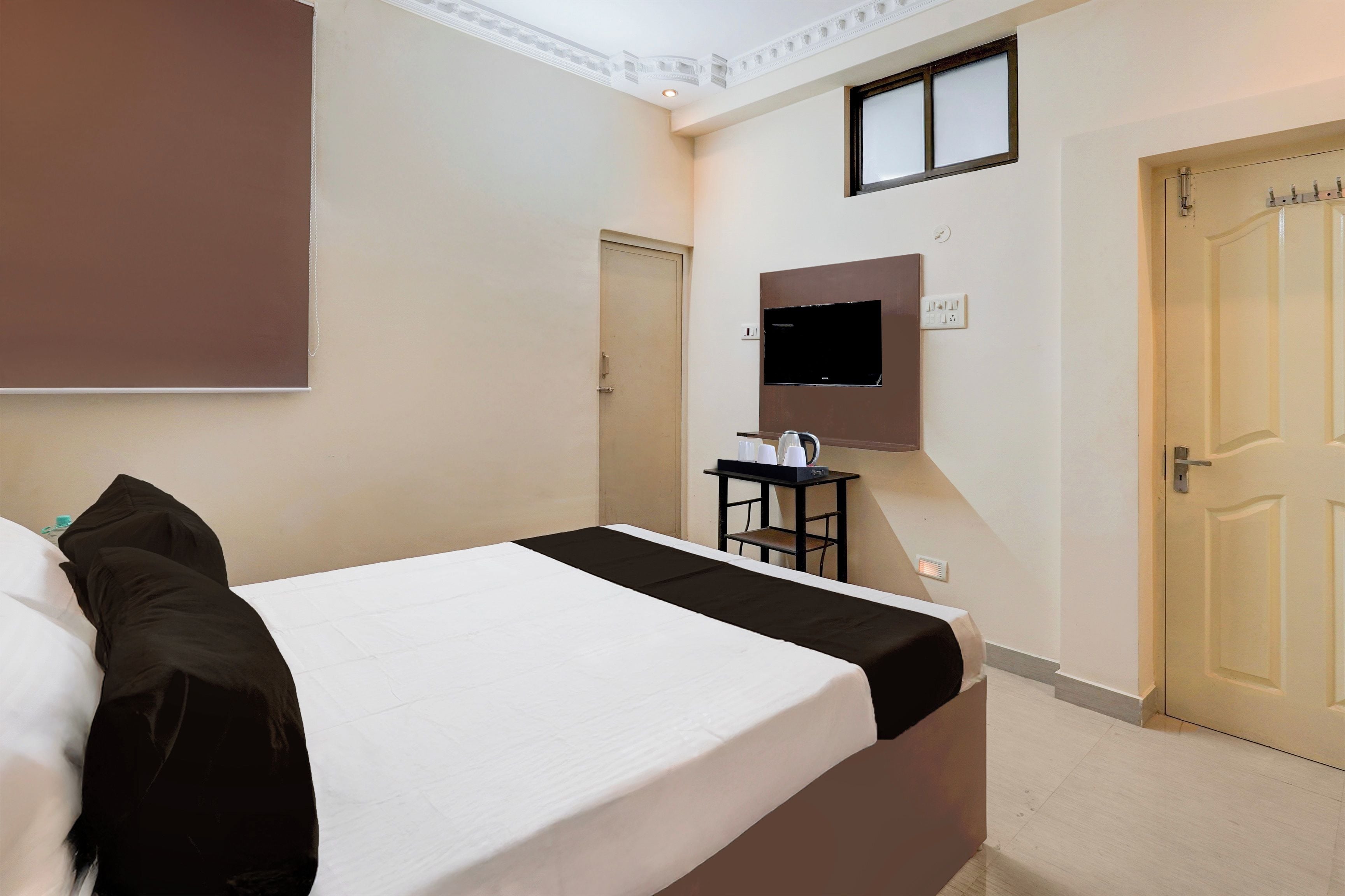 A hotel room with a bed, a television mounted on the wall, a small table with a kettle, and a door.