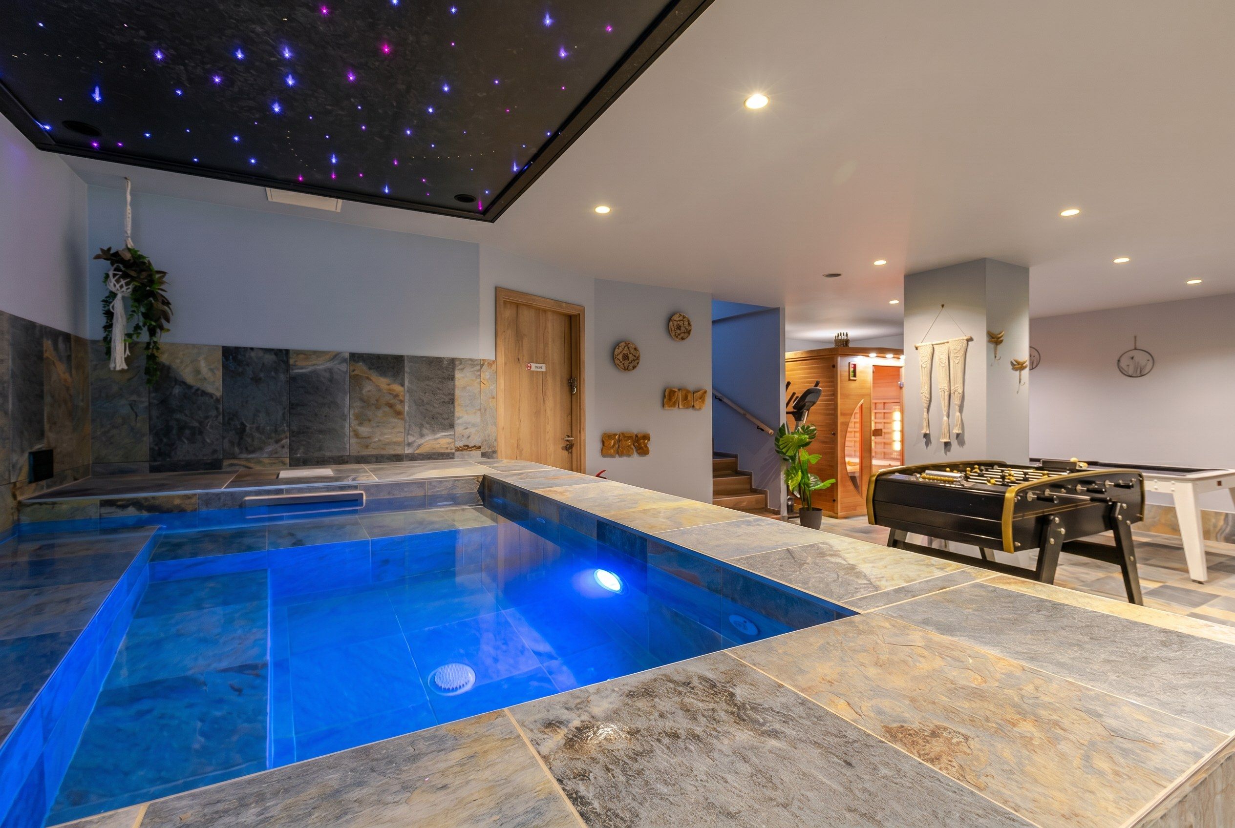 Un espace piscine intérieur moderne, avec une piscine à l’eau d’un bleu limpide, un sol en pierre et un plafond orné de projections d’étoiles.