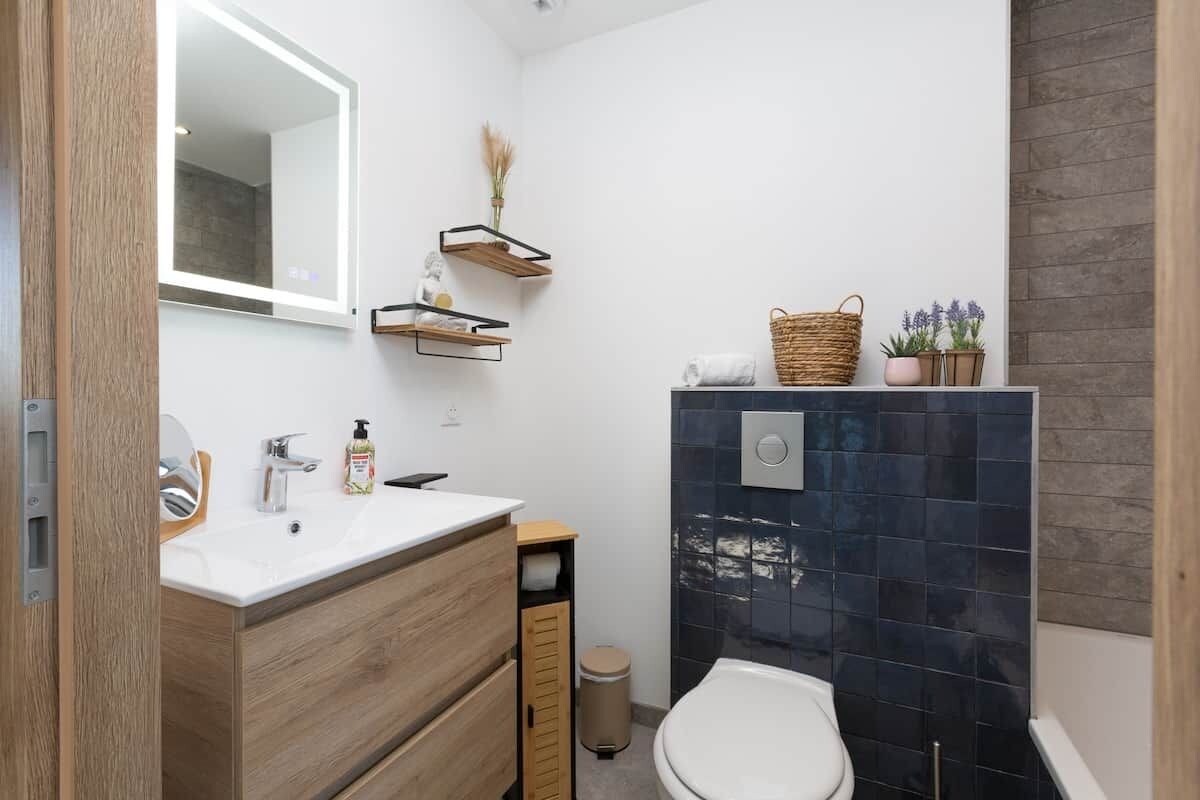 Une salle de bain moderne avec un lavabo blanc, un meuble-lavabo en bois, un miroir et une cabine de douche carrelée aux couleurs sombres.