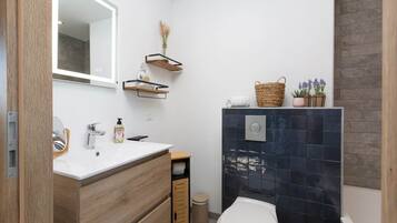 Une salle de bain moderne avec un lavabo blanc, un meuble-lavabo en bois, un miroir et une cabine de douche carrelée aux couleurs sombres.