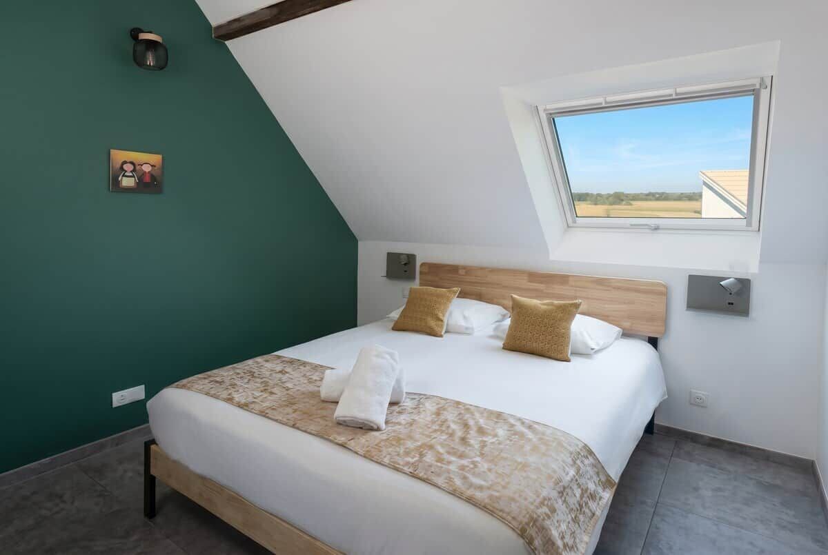 Una camera da letto con soffitto spiovente, un letto con lenzuola bianche, due cuscini, una lampada da comodino e un piccolo quadro appeso al muro.
