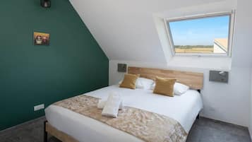 Una camera da letto con soffitto spiovente, un letto con lenzuola bianche, due cuscini, una lampada da comodino e un piccolo quadro appeso al muro.