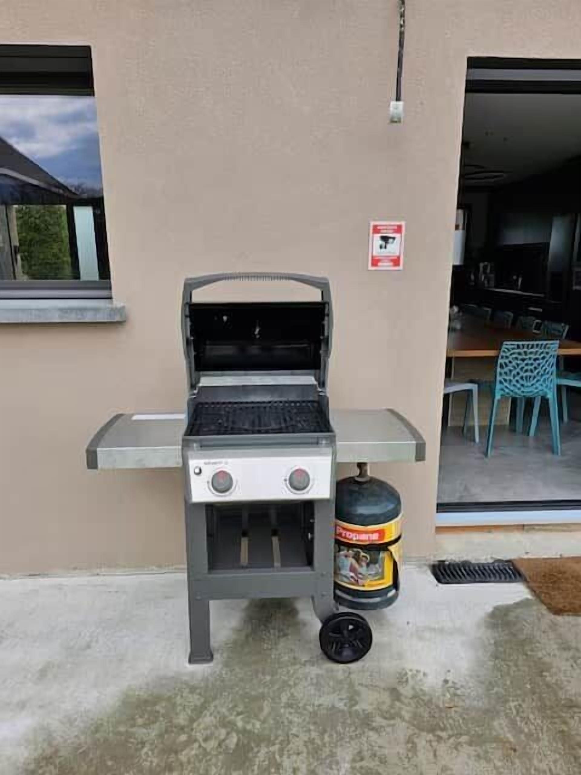 En udendørs grill med en propanflaske, et vindue og en dør, der fører til et indendørs område med stole.