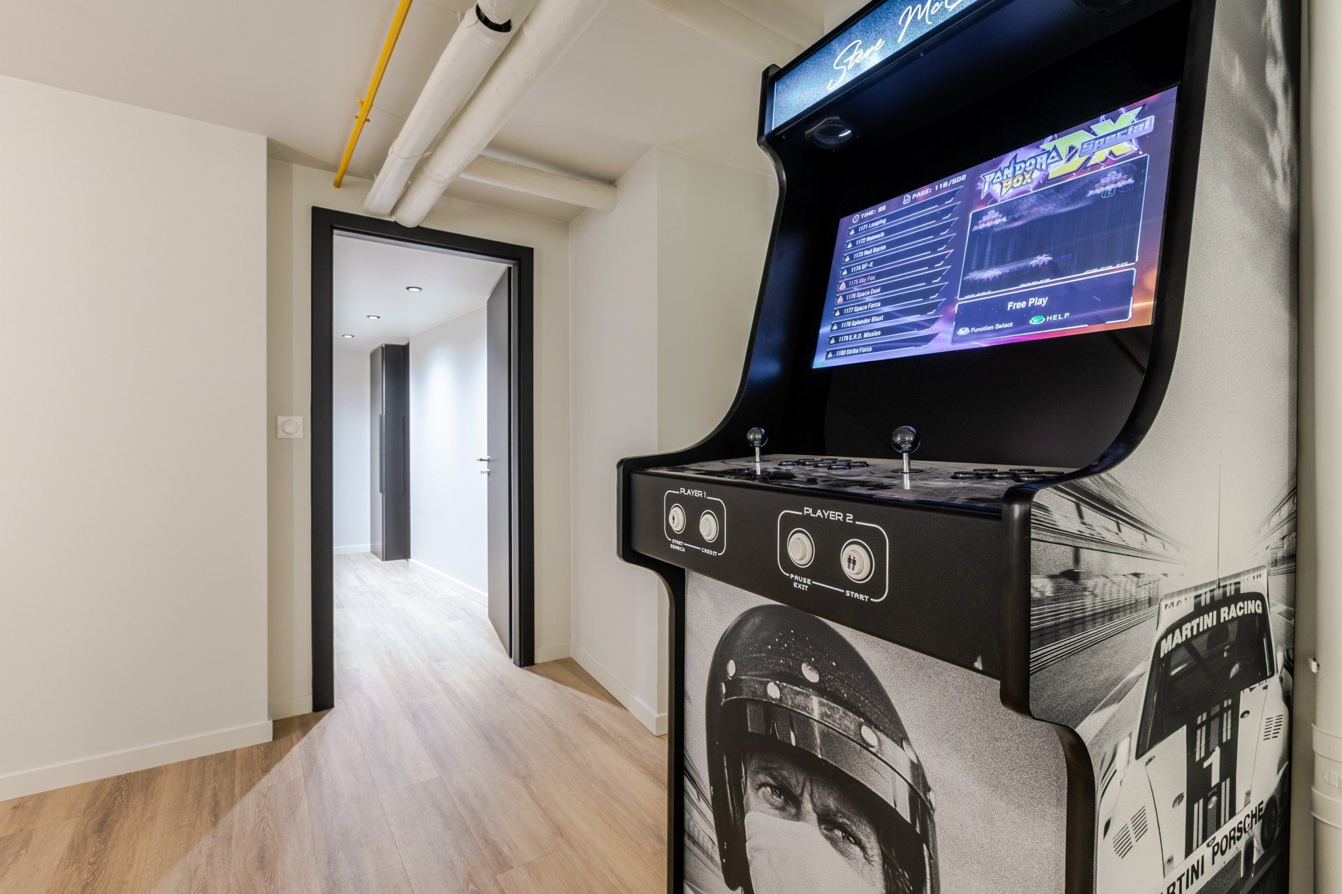 Een gameconsole met een beeldscherm waarop een racegame draait, in een hal met een houten vloer en een deur naar een andere kamer.