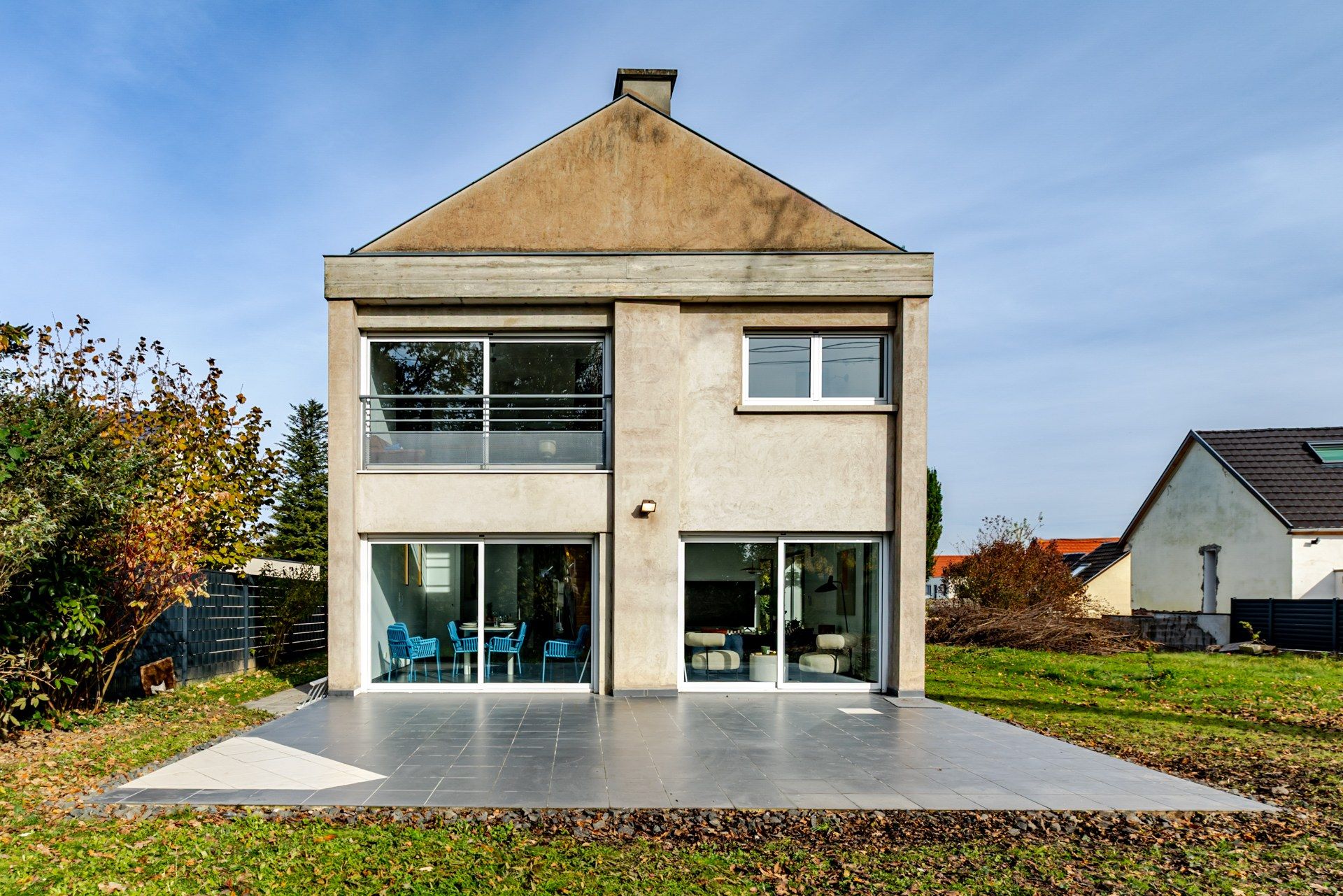 Une maison à deux étages, dotée d’un toit plat, de grandes fenêtres et d’un patio aménagé avec des sièges d’extérieur.