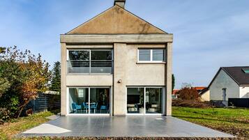 Une maison à deux étages, dotée d’un toit plat, de grandes fenêtres et d’un patio aménagé avec des sièges d’extérieur.