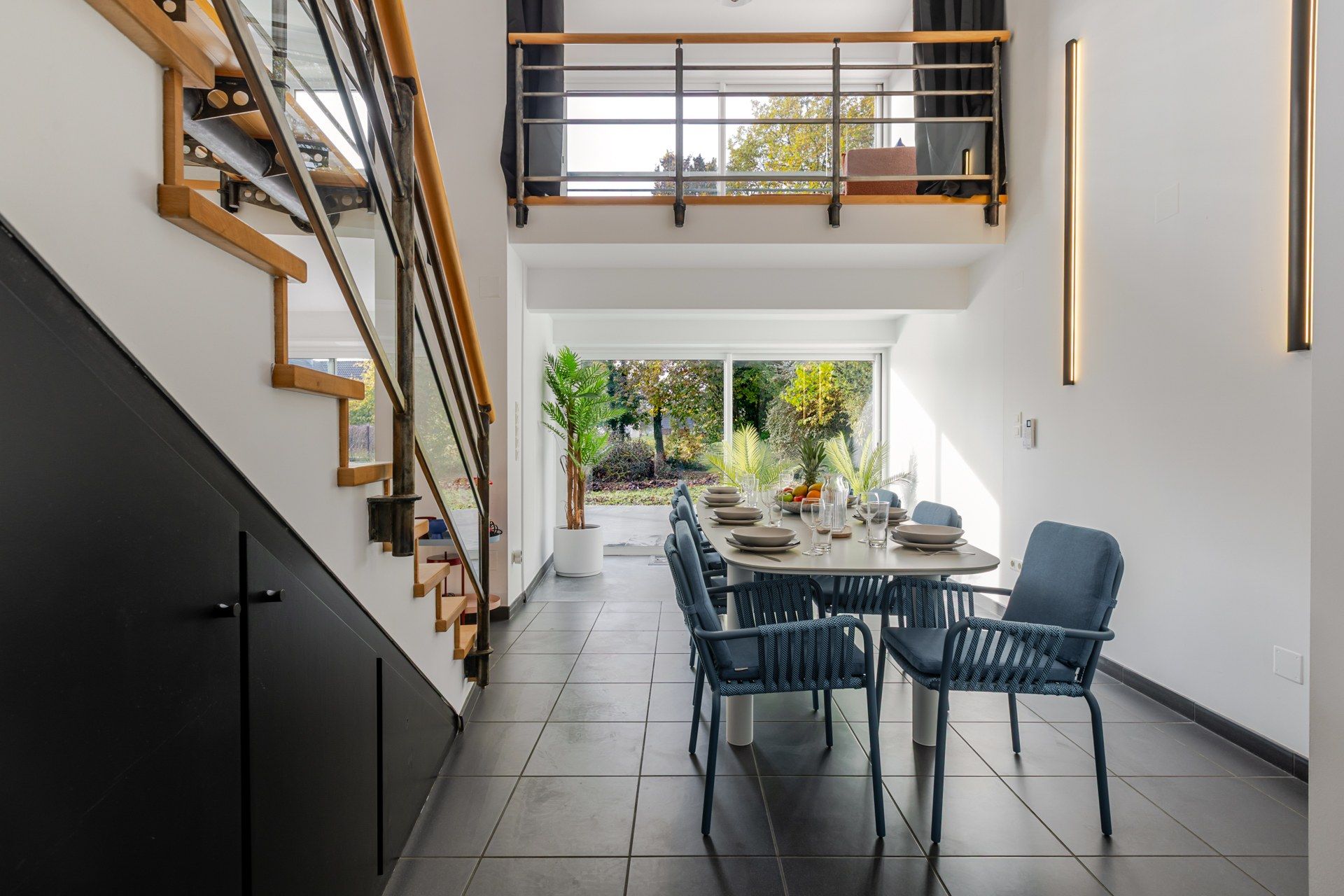 Un intérieur moderne avec un coin repas comprenant une table ronde et des chaises bleues, un escalier agrémenté d’éléments en bois et une vue sur l’extérieur.