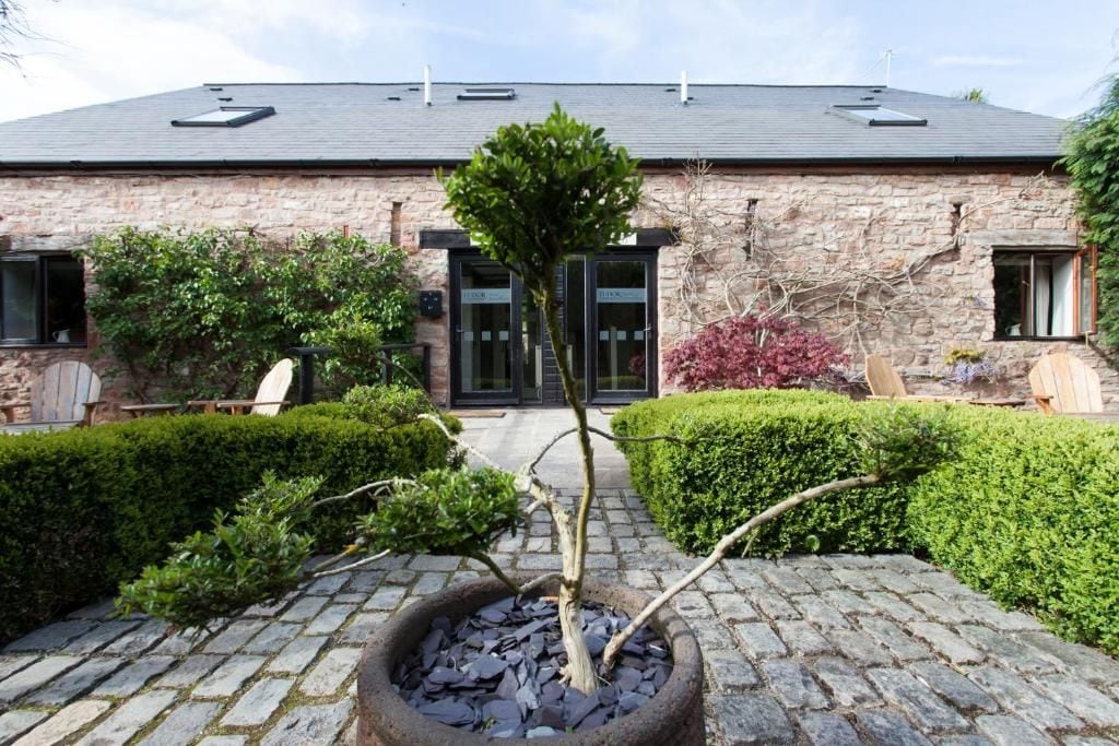 Un edificio de piedra con un sendero pavimentado, un árbol en maceta y setos bien cuidados.