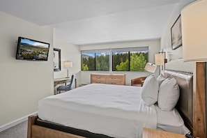 A hotel room with a large bed, a desk, a chair, a window with a forest view, and a television on the wall. - Beaver Run -Breck Blast|Ski Views|Pool&Hot Tubs (Breckenridge)