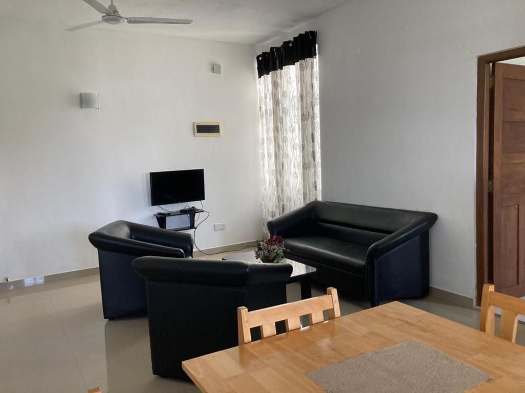 Una sala de estar con una mesa de comedor de madera, dos sofás negros, un televisor y una ventana con cortinas.