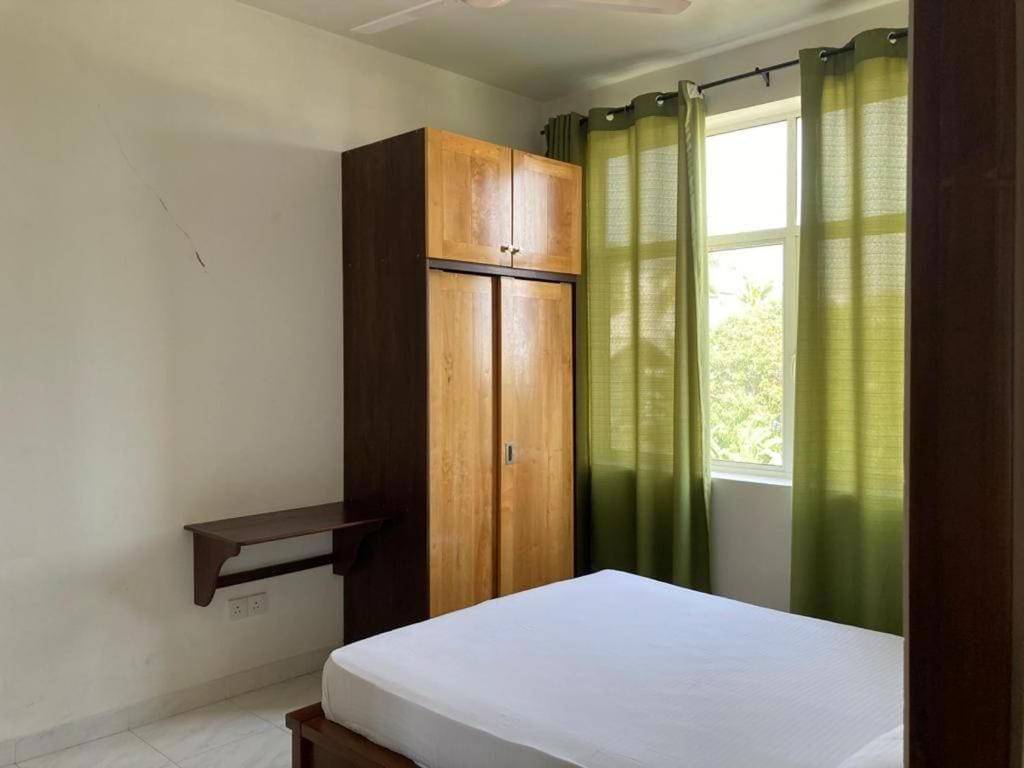 A bedroom with a bed, a wooden wardrobe, a shelf, and a window with curtains.