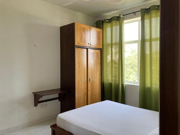 A bedroom with a bed, a wooden wardrobe, a shelf, and a window with curtains.