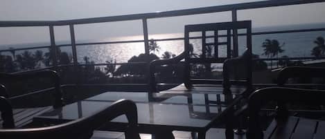 An outdoor seating area with tables and chairs, overlooking a body of water and palm trees.