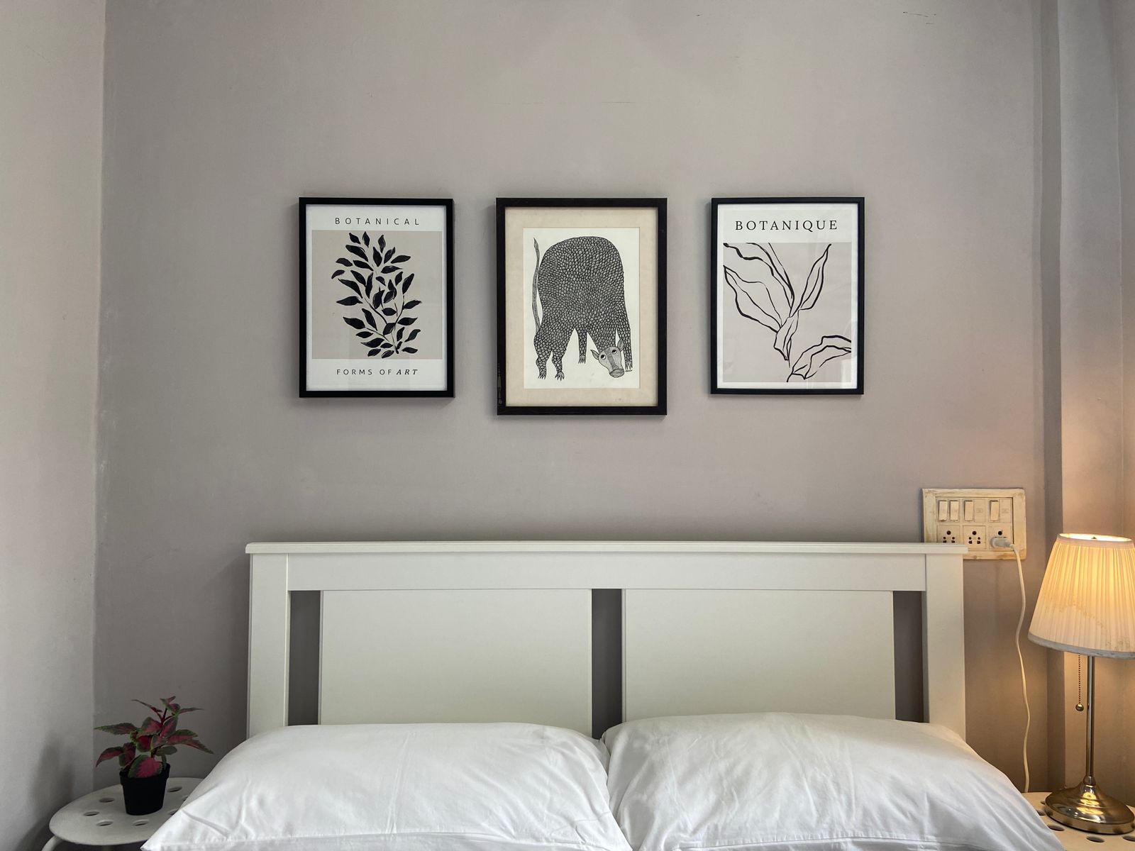 Une chambre à coucher avec un lit, une table de chevet avec une lampe et trois reproductions botaniques encadrées accrochées au mur.