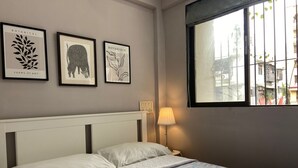 Una cama con sábanas blancas y una colcha azul estampada, una lámpara de noche y tres láminas enmarcadas con motivos botánicos en la pared.