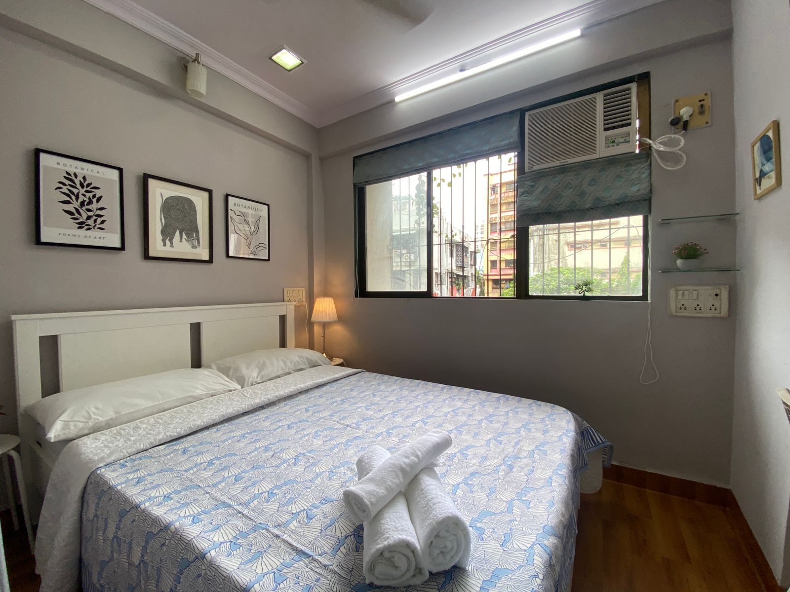 Une chambre d’hôtel avec un lit, une fenêtre donnant sur un beau paysage, un climatiseur fixé au mur et des œuvres d’art botanique encadrées.