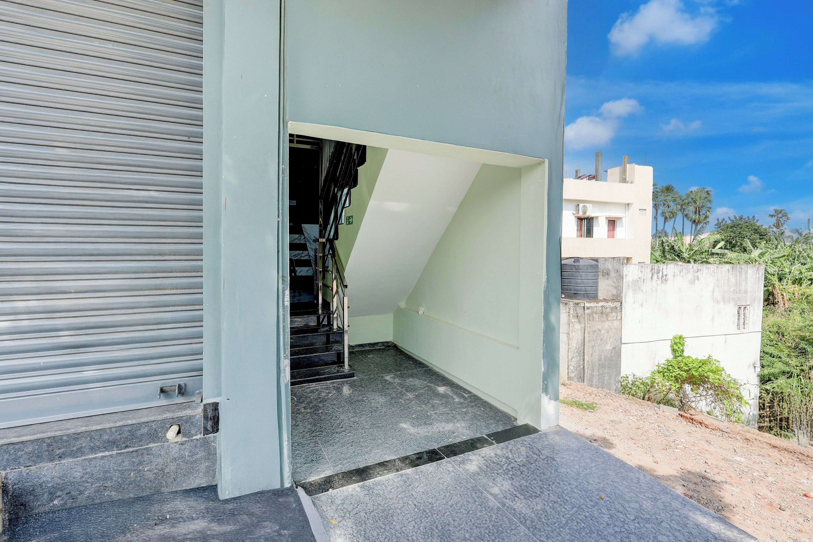 Entrada de un edificio con un cerrojo metálico cerrado, una escalera que da acceso al interior y vista de una zona residencial con casas y vegetación.
