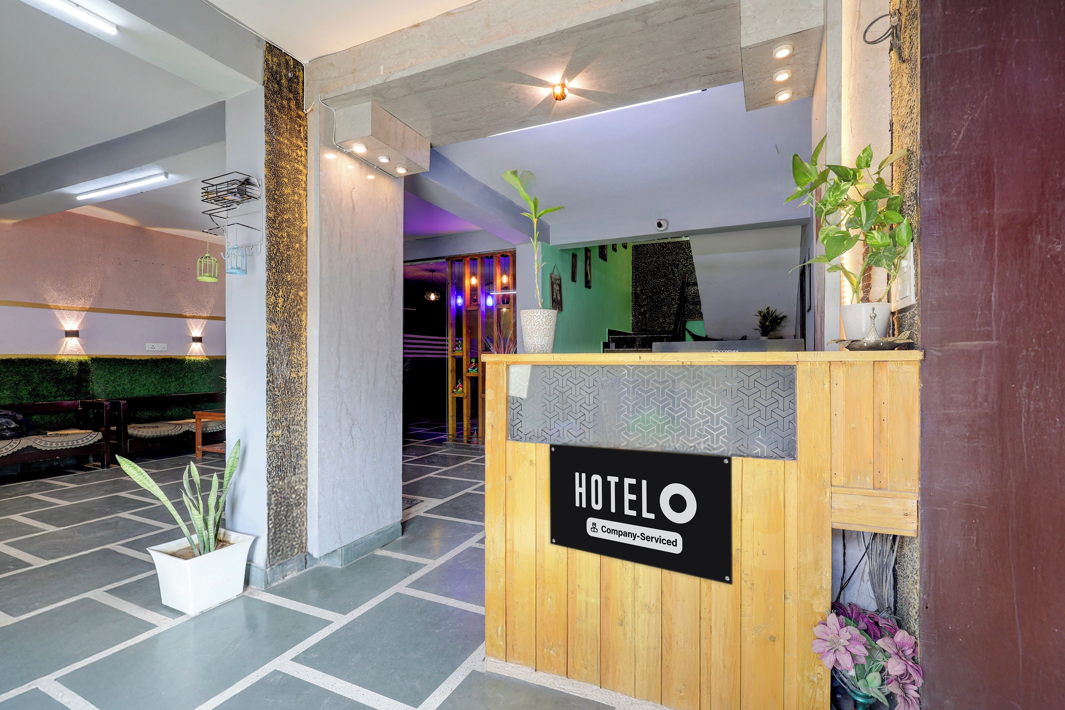 A hotel reception area with a wooden reception desk, a sign indicating it is company-serviced, and a green wall with a plant.