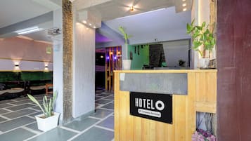 A hotel reception area with a wooden reception desk, a sign indicating it is company-serviced, and a green wall with a plant.