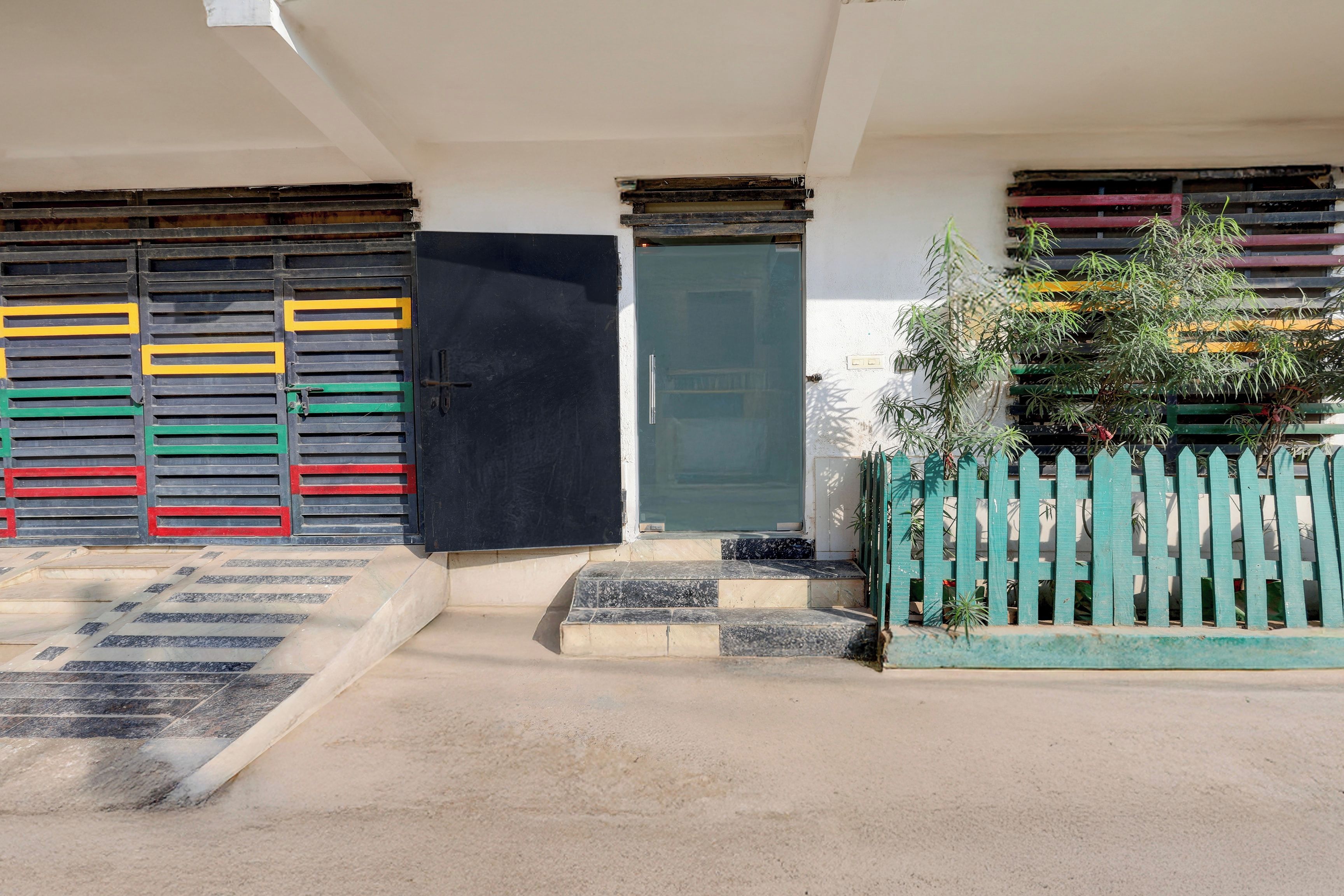 A building entrance with a set of stairs leading up to a door, a small garden area with a fence, and a wall with colorful shutters.