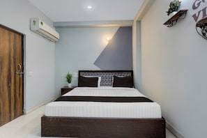 Quarto com uma cama, porta de madeira, ar condicionado, mesa de cabeceira com planta e prateleira na parede.