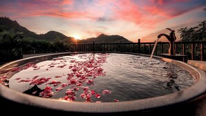 A circular hot tub with water and rose petals, surrounded by a railing with a view of mountains and a sunset. - Lamia Tropical Garden (Tay Hoa Lu)