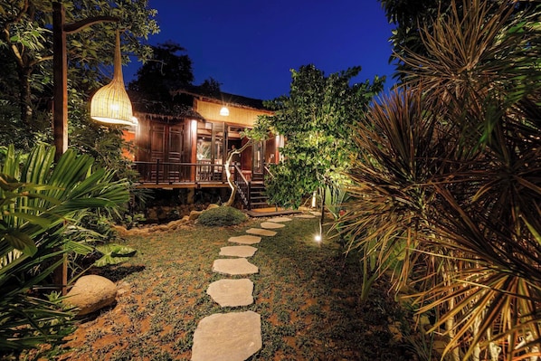 A well-lit pathway leading to a wooden house with a thatched roof, surrounded by tropical vegetation.