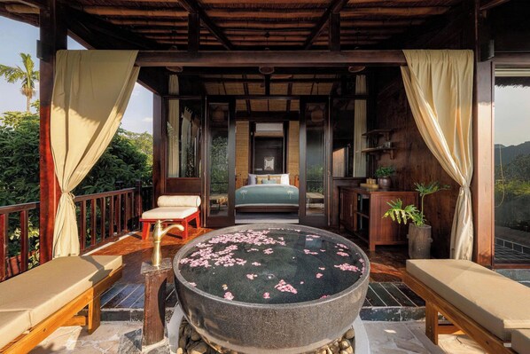 A bedroom with a bed, a small table, and a large stone basin with water and flower petals.