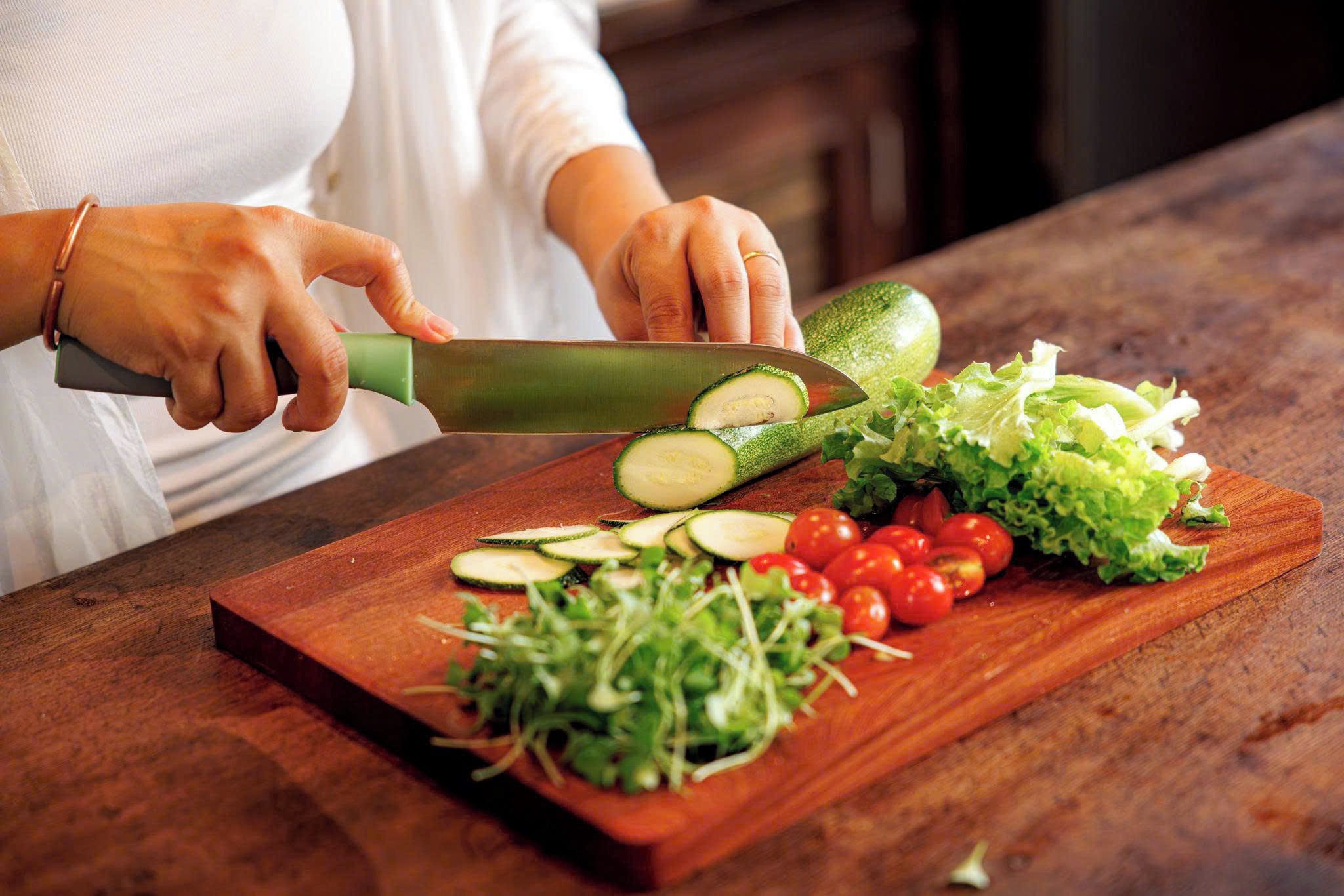 Une personne est en train de trancher un concombre sur une planche à découper en bois, avec d’autres légumes et des feuilles vertes à proximité.