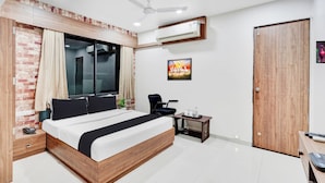 A modern bedroom with a wooden bed, a small bedside table, a desk with a chair, a window with curtains, a ceiling fan, and an air conditioning unit. - Collection Kudasan Gandhinagar Mansion (Gandhinagar)