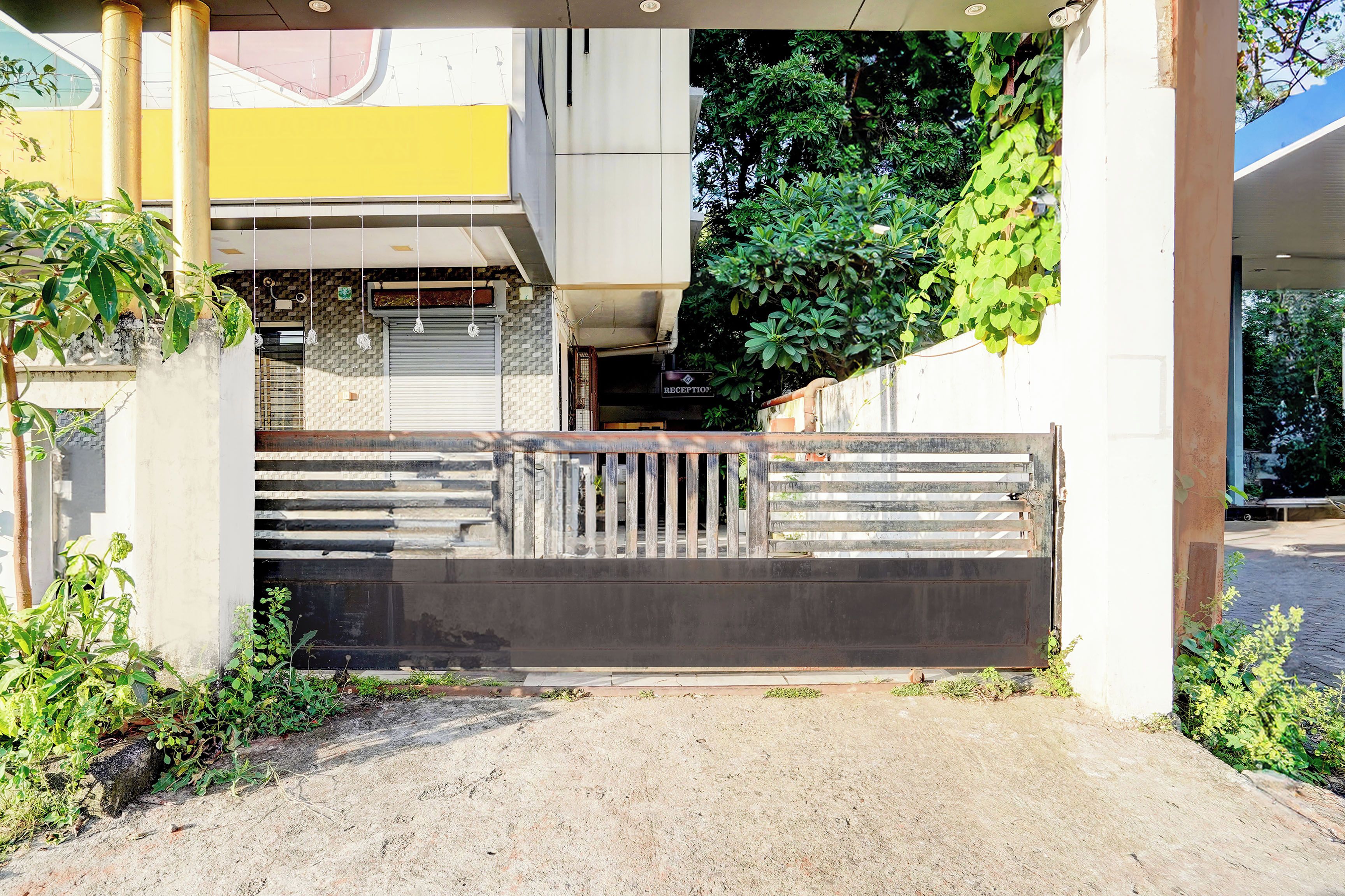 A gated entrance with a metal gate, concrete pillars, and a building with a yellow and white facade in the background.