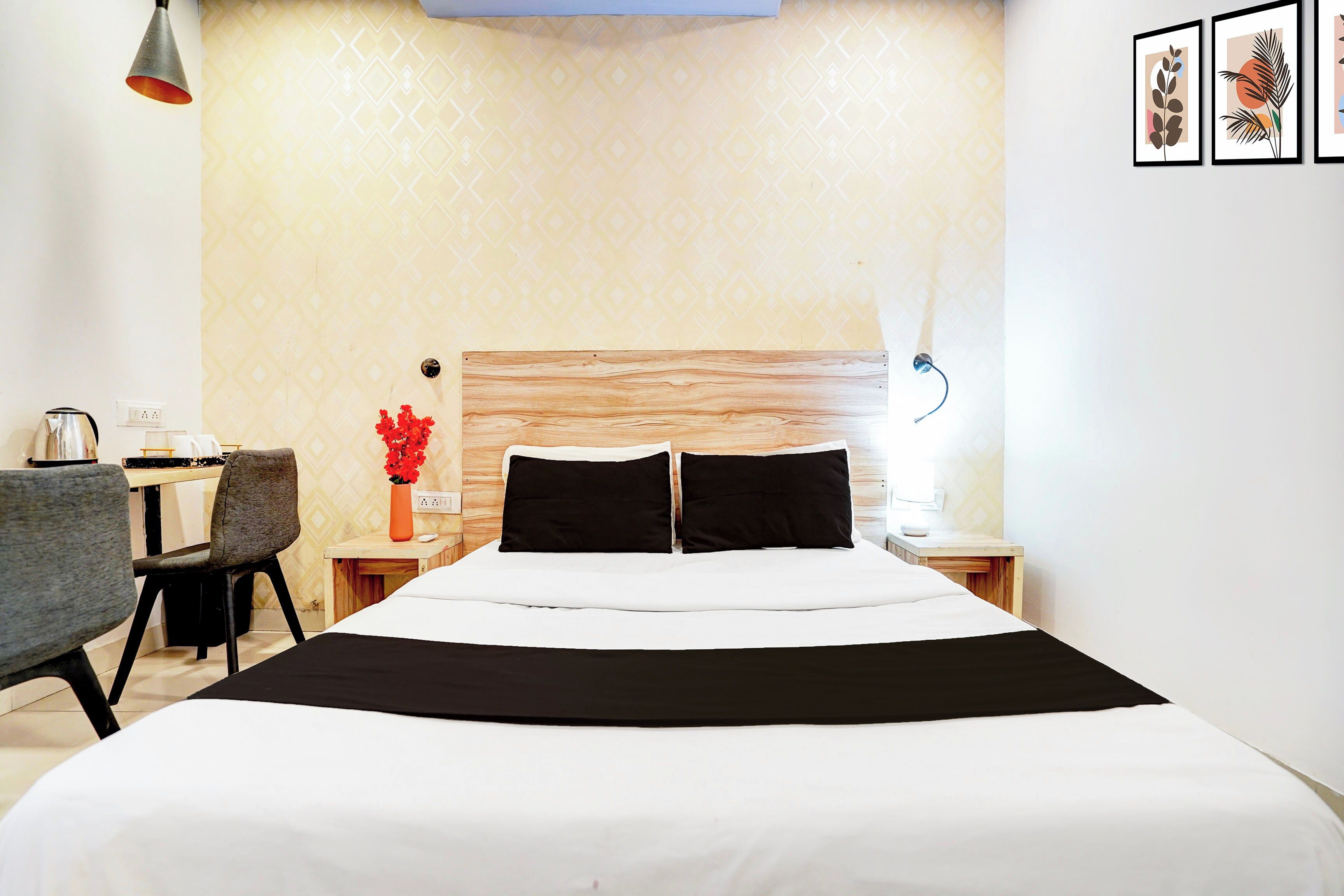 A hotel room with a bed, a desk, a chair, a nightstand, a vase with flowers, and framed artwork on the wall.