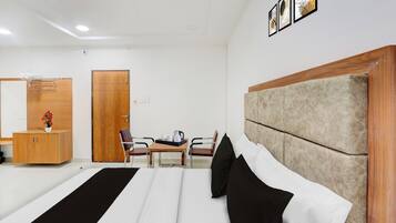 A hotel room with a bed, a wooden wardrobe, a desk with a chair, and a wall-mounted shelf.