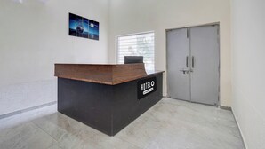 A reception desk with a sign indicating 'HOTEL' and 'Company Serviced.' There is a window with blinds and a door to the right.