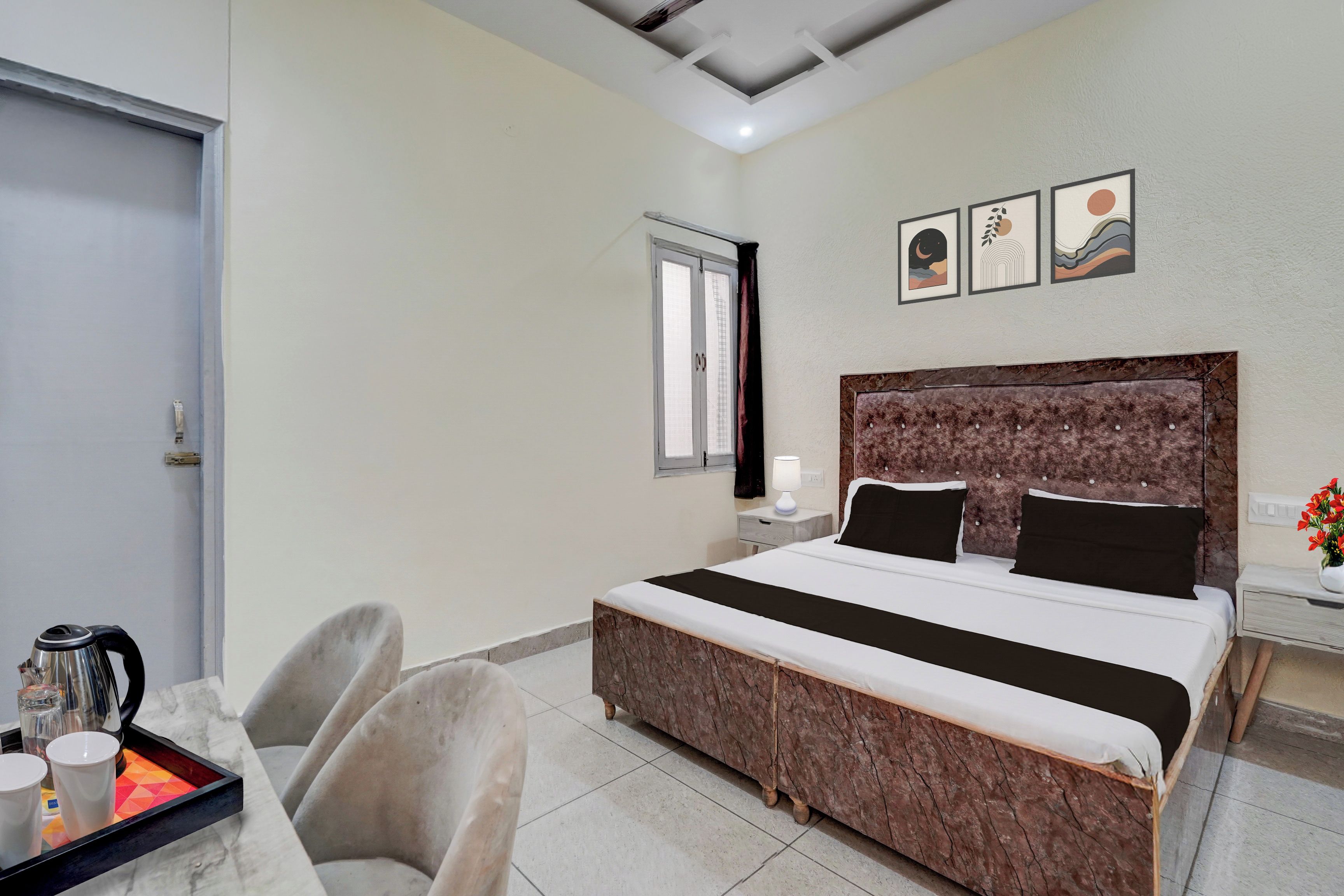 A hotel room with a bed, a desk with a kettle, chairs, a window, and artwork on the wall.