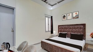 A hotel room with a bed, a desk with a kettle, chairs, a window, and artwork on the wall.
