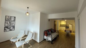Un salon comprenant un canapé, deux fauteuils, un coin repas avec une table et des chaises, une cuisine à l’arrière-plan et un luminaire au plafond.