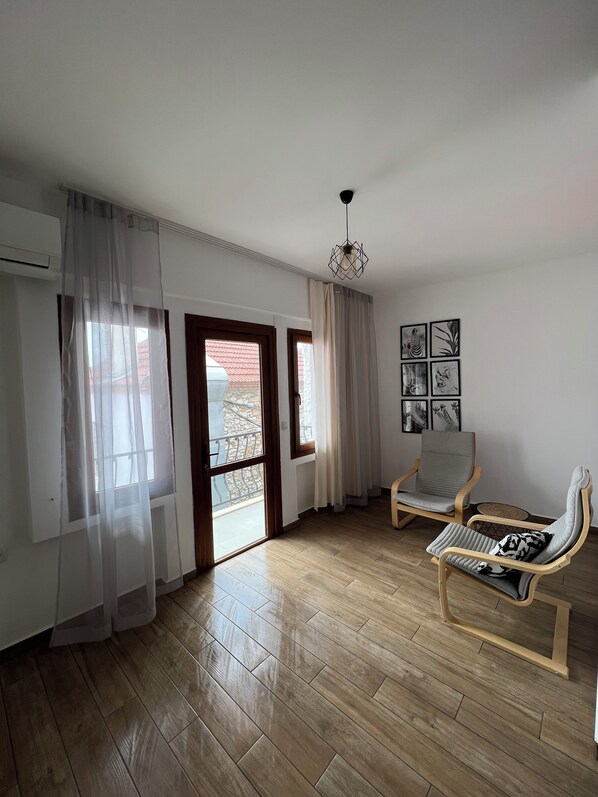 Une pièce avec un plancher en bois, une chaise, un canapé, un luminaire au plafond et une vue sur l’extérieur, visible à travers une porte.