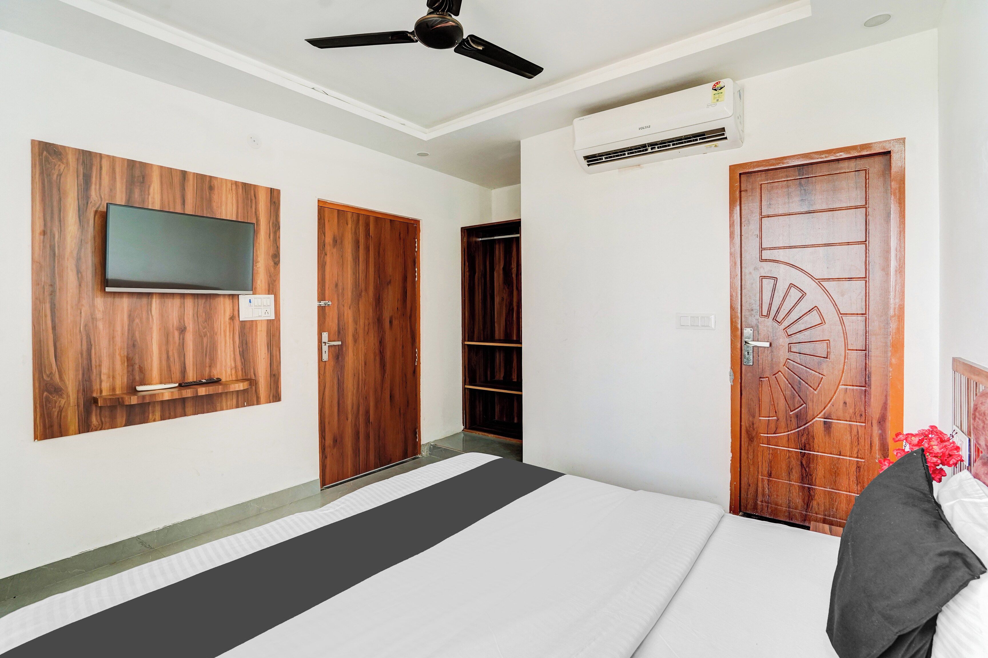 A hotel room with a bed, a flat-screen TV mounted on a wooden panel, a ceiling fan, and a door with a decorative design.
