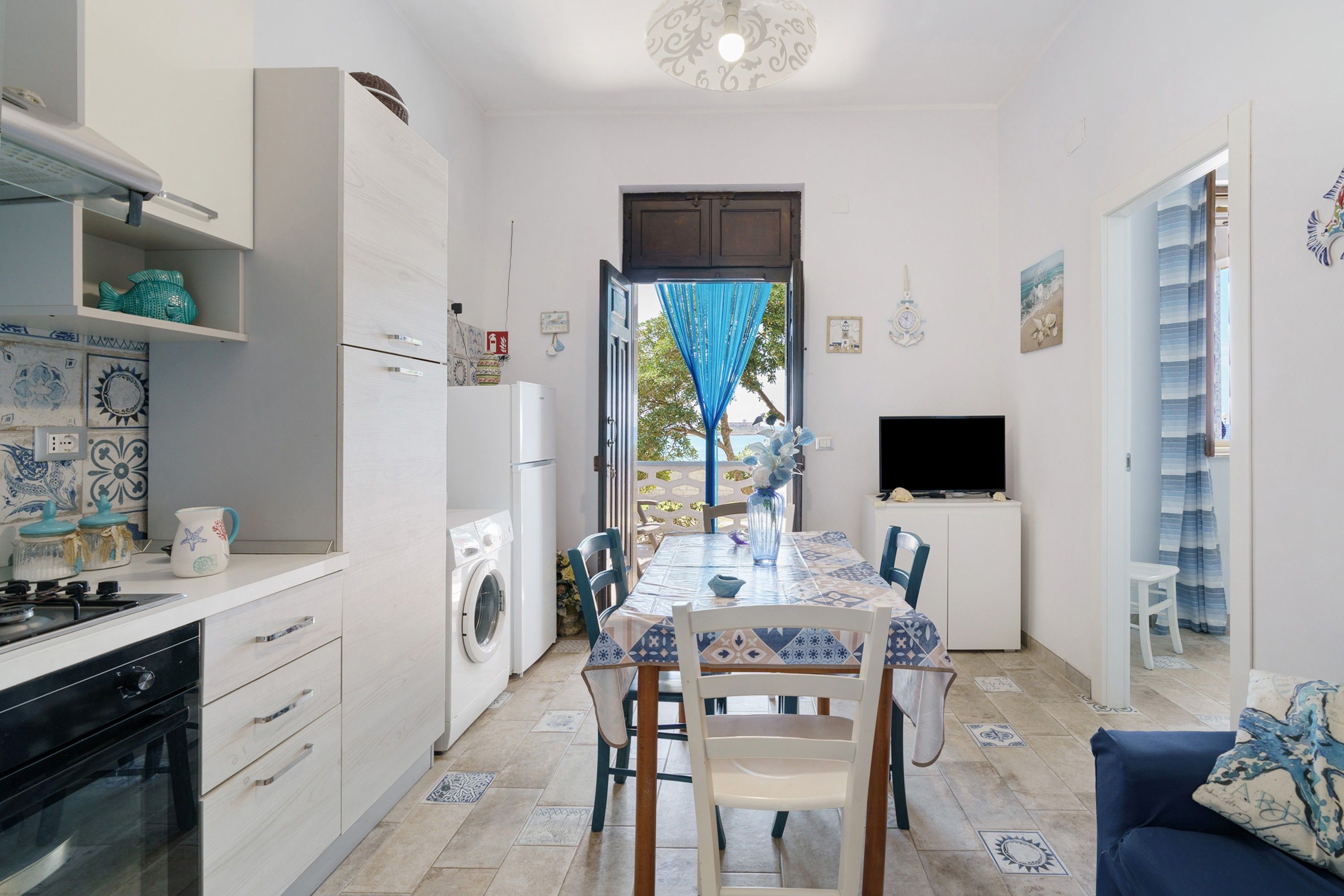 Een compacte keuken met een wasmachine, een eethoek met een tafel en stoelen, een televisie en uitzicht door een open deur.