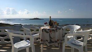 Une table dressée avec des chaises, donnant sur une vue de la côte, une bouteille et un panier de fruits.