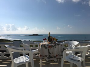 Une table dressée avec des chaises, donnant sur une vue de la côte, une bouteille et un panier de fruits.